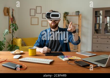Serious bearded designer sitting at table and drawing on digitizer while using virtual reality simulator at home Stock Photo