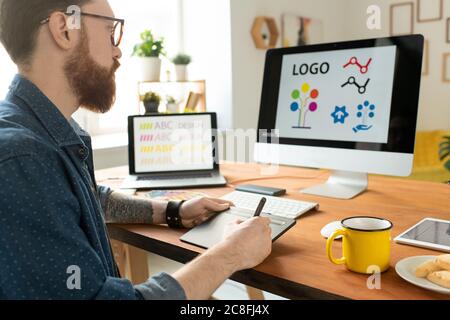 Over shoulder view of concentrated bearded designer in glasses sitting at desk and using drawing pad while working on website graphics Stock Photo