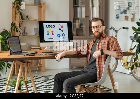 Portrait of serious handsome bearded brand designer sitting at desk with desktop computer and laptop in home office Stock Photo