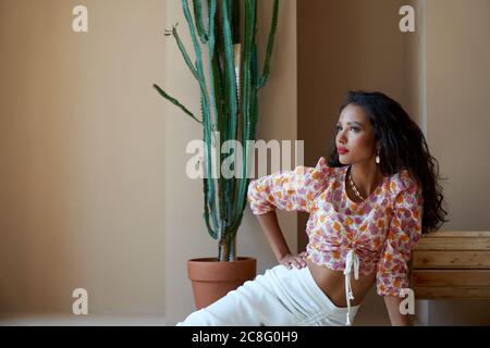 Portrait of mulatto girl with long black curly hair posing on floor near big cactus and wooden wheel. Stunning half african brunette wearing blouse with floral print, white skirt and seashells. Stock Photo