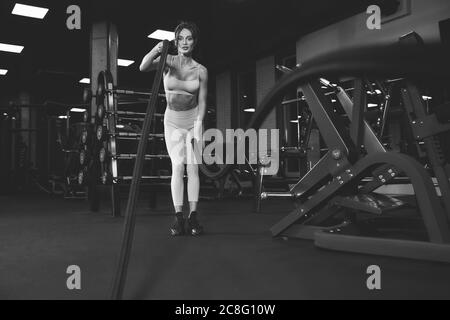 From below view of young woman in light sportswear training with ropes. Monochrome portrait of attractive brunette fitnesswoman with strong face doing cardio in empty gym. Concept of sport. Stock Photo