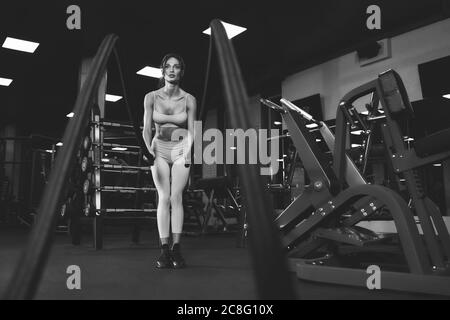 From below view of brunette woman in light sportswear training in empty gym with ropes. Monochrome full length portrait of attractive fitnesswoman with strong face doing cardio. Concept of sport. Stock Photo