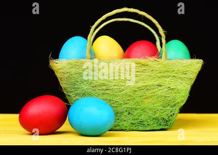 Basket full of colourful painted eggs for Easter and several other bicolor eggs lying on wooden table isolated on black background, close up. Easter collection concept Stock Photo