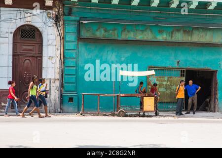 Havana / Cuba - 04.15.2015: Three Cuban women walking in front of a work place where two Cuban men are standing and talking, two kids playing on an ol Stock Photo