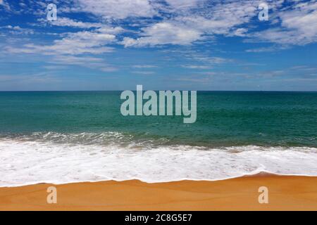 Surf on a tropical sandy beach, scenic view to empty sea coast with yellow sand and emerald wave with white foam. Picturesque seascape with blue sky Stock Photo