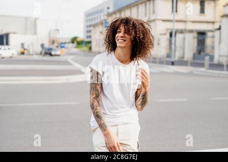 Smiling man with tattooed arms and long brown curly hair standing on a street. Stock Photo