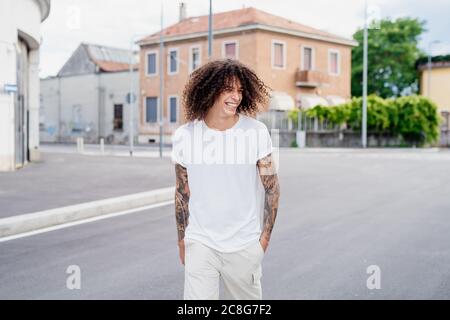 Smiling man with tattooed arms and long brown curly hair walking down street. Stock Photo