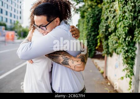 Two men with tattooed arms standing on sidewalk, hugging. Stock Photo