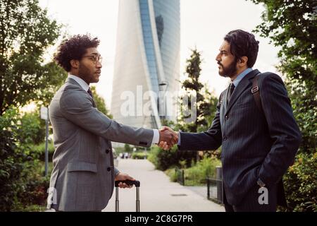 Two businessmen wearing suits standing outdoors, shaking hands. Stock Photo