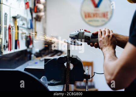 Young man using angle grinder on metal in workshop, cropped Stock Photo