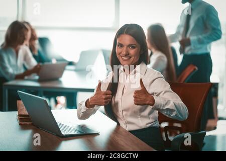 Businesswoman shows thumbs up in office. Alright gesture from young smiling girl. Group of businesspeople work on blurred background. Achievement in Stock Photo