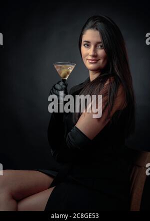 Attractive girl drinking dry martinis with green olives. Beautiful brunette in black holding glass with cold cocktail. Stock Photo