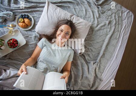 attractive woman lies on bed in the morning, drinking coffee, reading book, casual style, grey dress, feeling comfortable at home, having rest Stock Photo