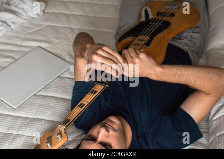 Man lying on a bed using a mobile phone after playing electric bass. Stock Photo
