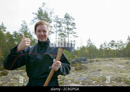 Portrait of happy young handsome man giving thumbs up while holding ax in the forest Stock Photo