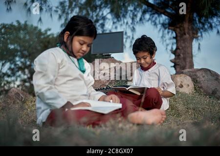 Two happy elementary students with a book and computer studying at natural park, New Normal Education concept Stock Photo