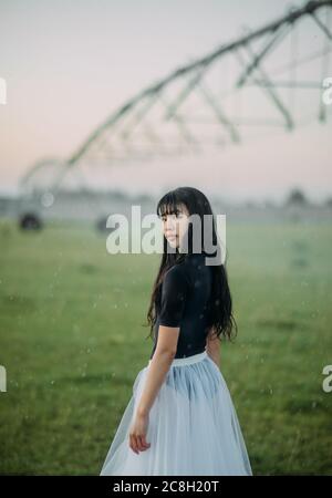 Japanese ballerina in white skirt stands in field on farm on background of agricultural sprayer. Stock Photo