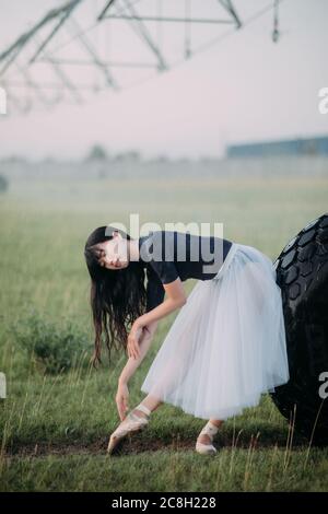 Japanese ballerina in white skirt dances in field on farm near the wheel of agricultural sprayer. Stock Photo