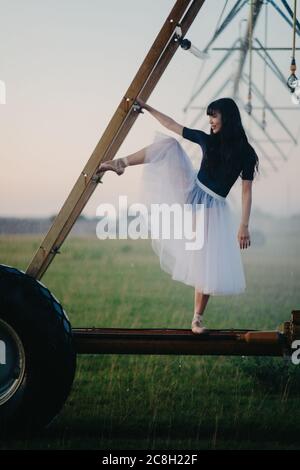 Japanese ballerina in white skirt dances in field on farm near the wheel of agricultural sprayer. Stock Photo