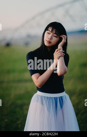 Japanese ballerina in white skirt stands in field on farm on background of agricultural sprayer. Stock Photo