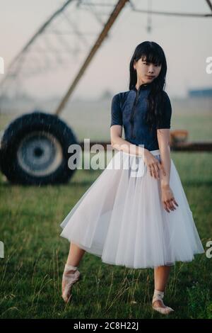 Japanese ballerina in white skirt dances in field on farm on background of agricultural sprayer. Stock Photo