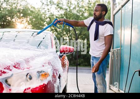 Portrait of handsome bearded young African man washing his red car with foam at at self service carwash outdoors. Clean car concept. Car wash Stock Photo