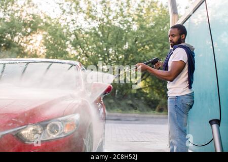 Handsome African young man in casual wear, washing his red car manually with water high-pressure hose at outdoor self wash service. Car washing Stock Photo