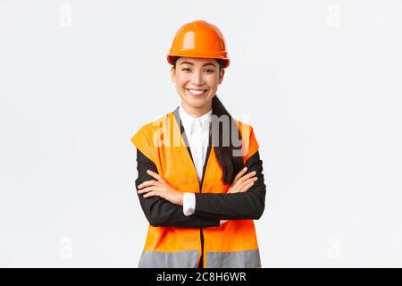 Smiling confident asian female chief manager at building, construction worker in safety helmet cross arms and looking determined. Assertive architect Stock Photo