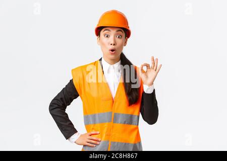 Imressed and surprised asian female construction manager pleased with amazing quality, standing in safety helmet and reflective jacket, showing okay Stock Photo
