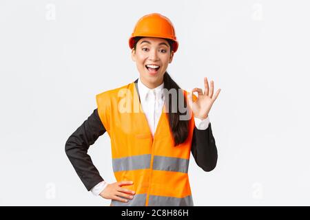 Satisfied happy asian female engineer, construction manager in safety helmet and reflective jacket showing okay gesture pleased, guarantee building Stock Photo