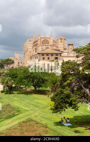 A couple sit in the shade of a tree Parc de la Mar with the Palma cathedral in the background, Mallorca, Spain Stock Photo