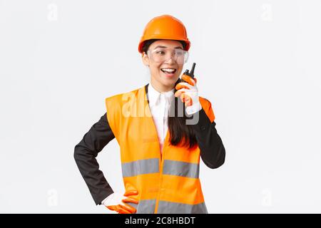Confident pleased asian female engineer in safety helmet and uniform talking with chief architect using walkie-talkie. Satisfied construction Stock Photo