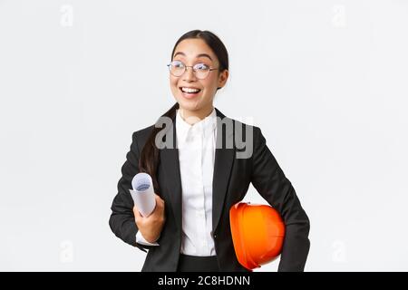 Hopeful and excited asian female architect looking around with fascination and amazement, holding blueprints and helmet, walking in construction area Stock Photo