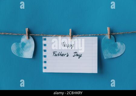 White notebook in line paper greeting card with black handwritten lettering happy fathers day hanging on brown rope with clothespins isolated on blue Stock Photo