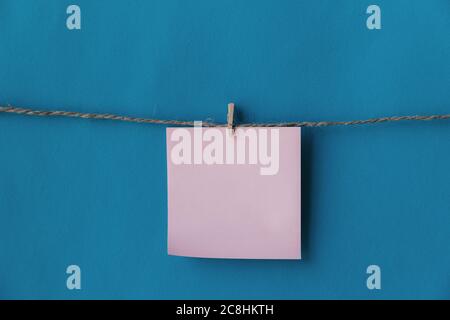 One pink sheet of paper with place for text hanging on rope isolated blue background. To do list. Pink sticky notes with cloth pegs. Pink notes paper. Stock Photo