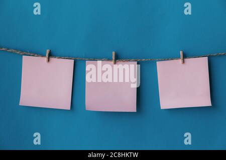 Three pink sheets of paper with place for text hanging on rope isolated blue background. To do list. Pink sticky notes with cloth pegs. Pink notes pap Stock Photo
