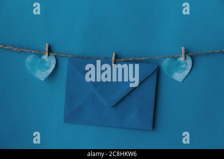 One blue envelope hanging on rope isolated blue background with two blue hearts on the sides. Write a letter to a friend. Postal envelope. Gift envelo Stock Photo