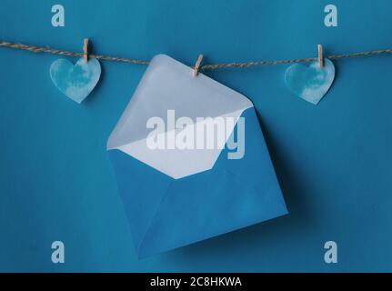 One opened blue envelope hanging on rope isolated blue background with two blue hearts on sides. Write a letter to a friend. Postal envelope. Gift env Stock Photo