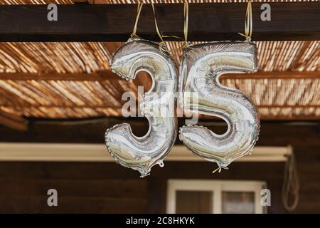 Happy 35th birthday party celebration balloon. Silver number thirty five 35 made of inflatable balloon with golden ribbon isolated on blurred brown ba Stock Photo