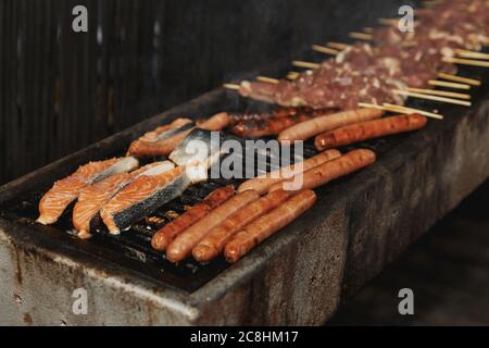 Close-up view of Grilling Chicken Meat, fish salmon steak and Sausages on long rectangular barbecue with smoke on the street outdoor. Summer barbecue Stock Photo