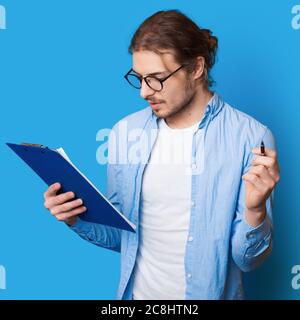 Bearded caucasian man with long hair making notes in a document holding a pen and folder on a blue studio wall Stock Photo