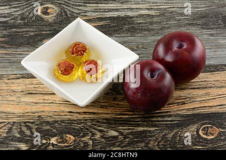 Plum hard candy drops with plum pit in center in white candy dish and fresh raw plum fruit on table Stock Photo