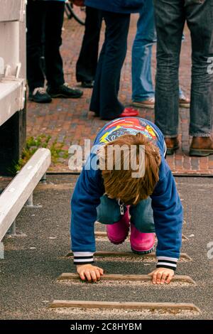 A small child is clinging on the wooden steps of a mild slope while adults are standing behind. this hyperactive child is bored being among adults and Stock Photo
