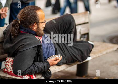 Paris, France 06/12/2010: Isolated image of a young homeless man wearing a hoodie and track pants as he is lying on his elbow on a wooden bench. He is Stock Photo