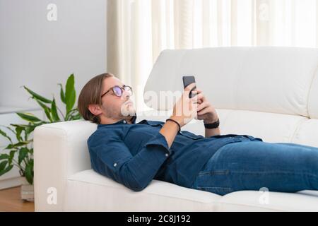Close up of a handsome young man lying on a sofa and focused on his phone on an out of focus background. Communication and leisure concept. Stock Photo