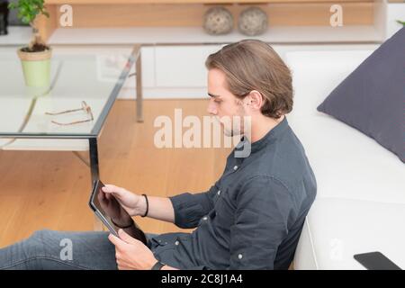 Side view of a handsome young man sitting on the ground next to a glass table checking a tablet on an out of focus background. Leisure and technology Stock Photo