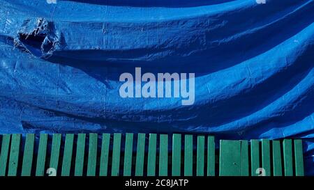 Folds of rumpled plastic construction fabric with torn hole and green painted wooden fence. Industrial blue green background with copy space Stock Photo