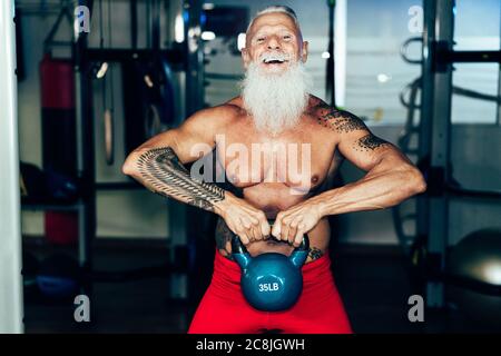 Hipster senior man training inside gym - Mature tattooed person having fun doing workout exercises in sport fitness club - Active joyful elderly lifes Stock Photo