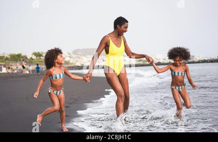 African sisters twins running on the beach with smiling mother - Black family people having fun on summer time - Vacation, travel and happiness lifest