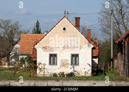 Abandoned small old red bricks suburban family house with cracked facade and two dilapidated wooden frame windows surrounded with other buildings Stock Photo